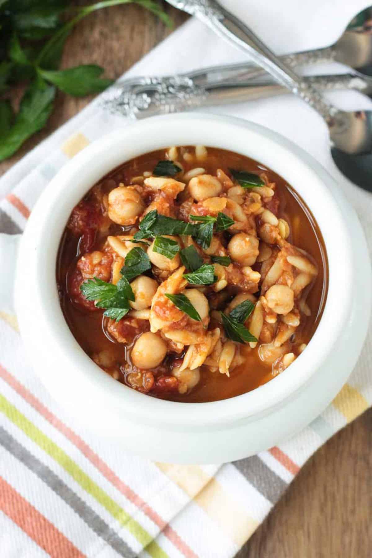 Red Lentil Stew with Chickpeas and Orzo is just as hearty as it's name. Luckily, you can throw all the ingredients in your slow cooker for a few hours and dinner is served! Healthy and nutritious and even my kids love this meal! A filling vegetarian dinner for Meatless Monday. Overhead view of red lentil stew in a white bowl garnished with parsley.