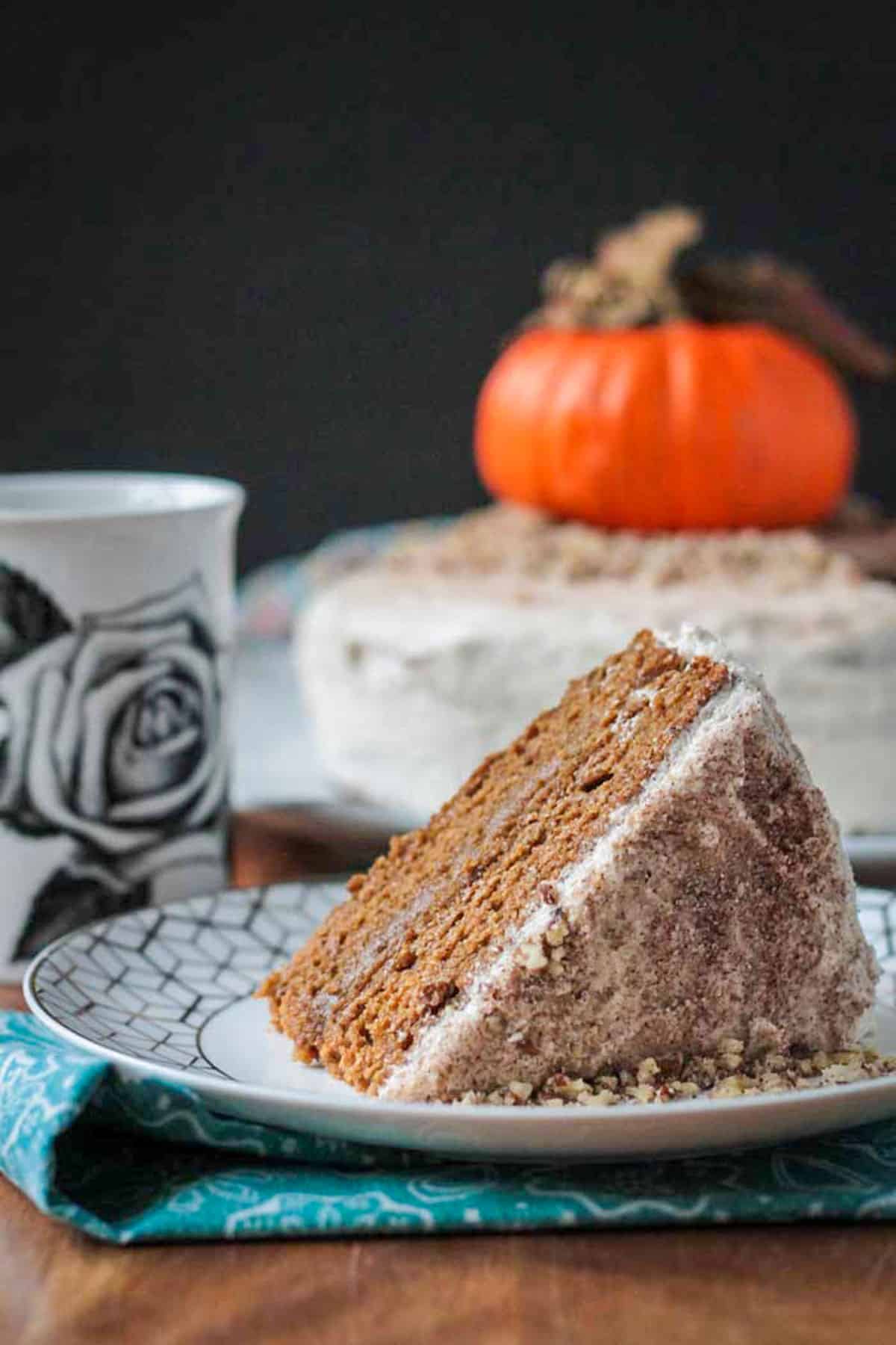 Pumpkin Layer Cake with chai spices, a thin layer of pumpkin cashew filling, and topped off with creamy dairy free cinnamon buttercream frosting. You will love this soft, moist vegan cake. Perfect for Halloween, Thanksgiving, or any fall entertaining. Slice of pumpkin layer cake on its side on plate with the full cake behind.