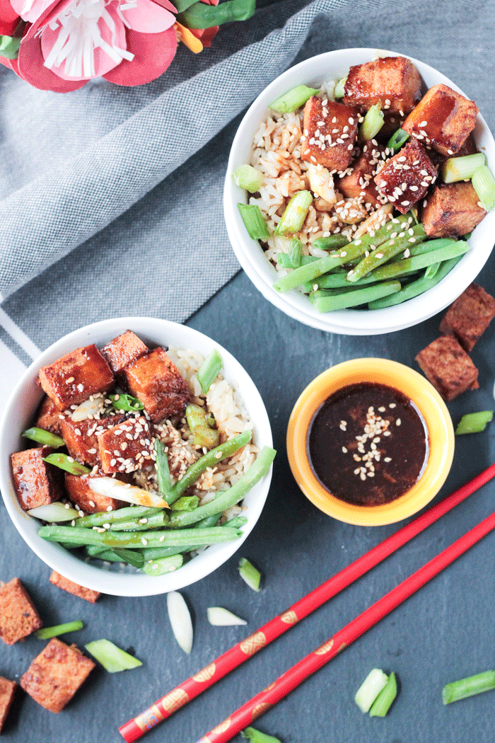 Two bowls of Sesame Ginger Baked Tofu over rice with green beans. Small yellow bowl of brown sauce next to the dinner bowls. Two red chopsticks in front. Gray dish cloth behind.