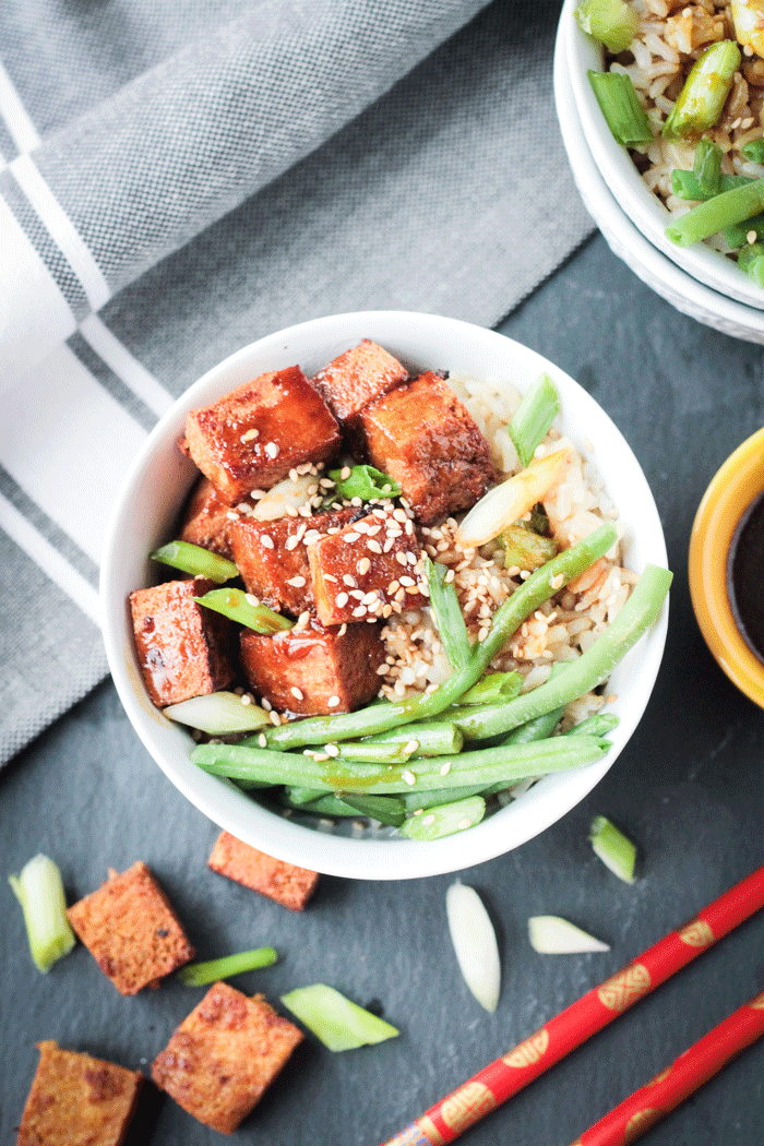 Bowl of Sesame Ginger Baked Tofu with greens beans over rice and sprinkled with sesame seeds. Gray dish cloth behind.