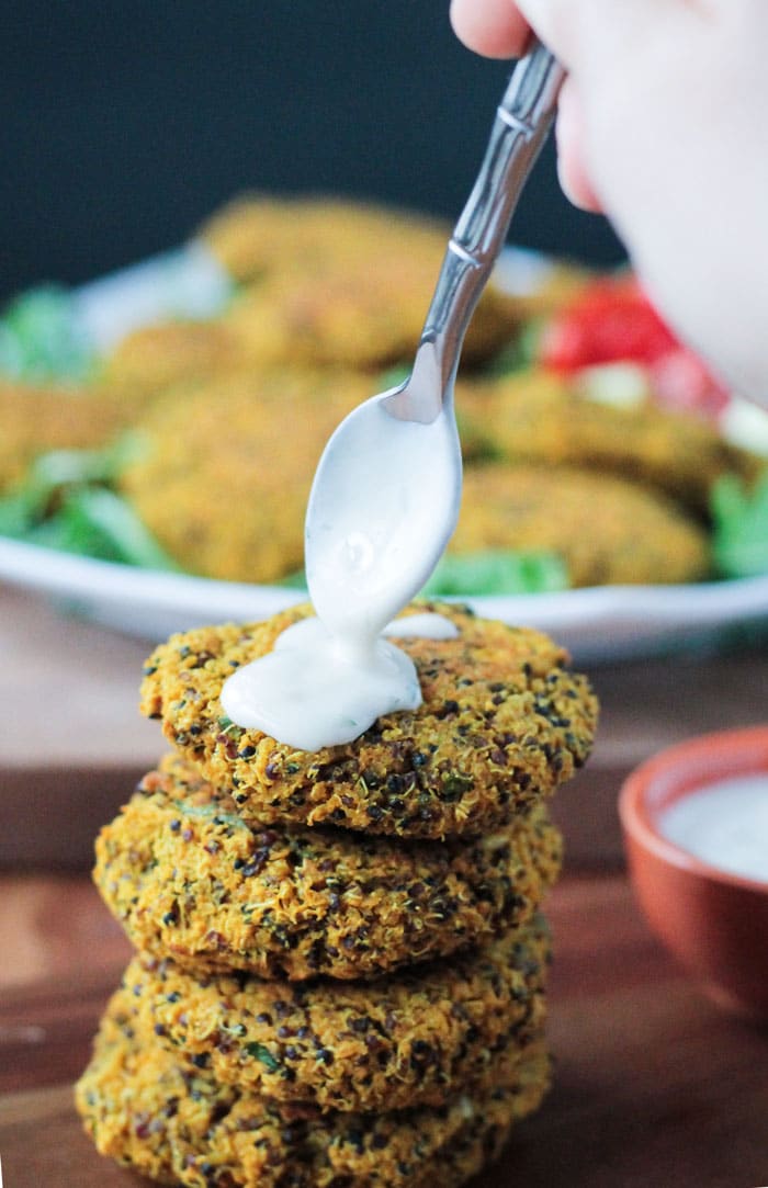 A spoon drizzling tahini yogurt sauce over a stack of four Baked Red Lentil Quinoa Fritters. Plate of fritters in the background.