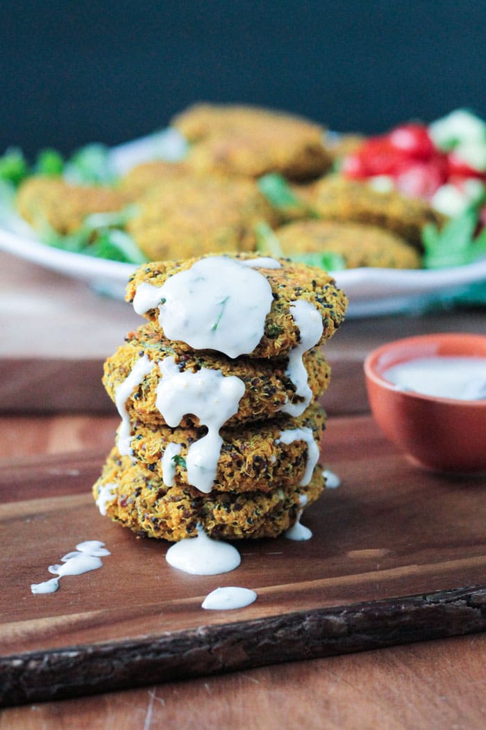Stack of Baked Red Lentil Quinoa Fritters drizzled with yogurt sauce. Small bowl of sauce nearby.