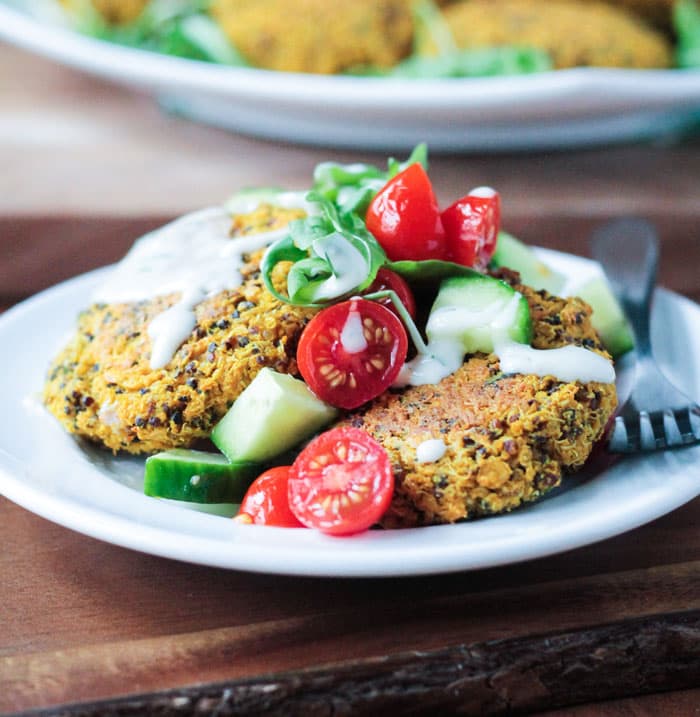 Plate of Baked Red Lentil Quinoa Fritters topped with yogurt sauce, tomato cucumber salad, and arugula. A fork rests on the side of the plate.