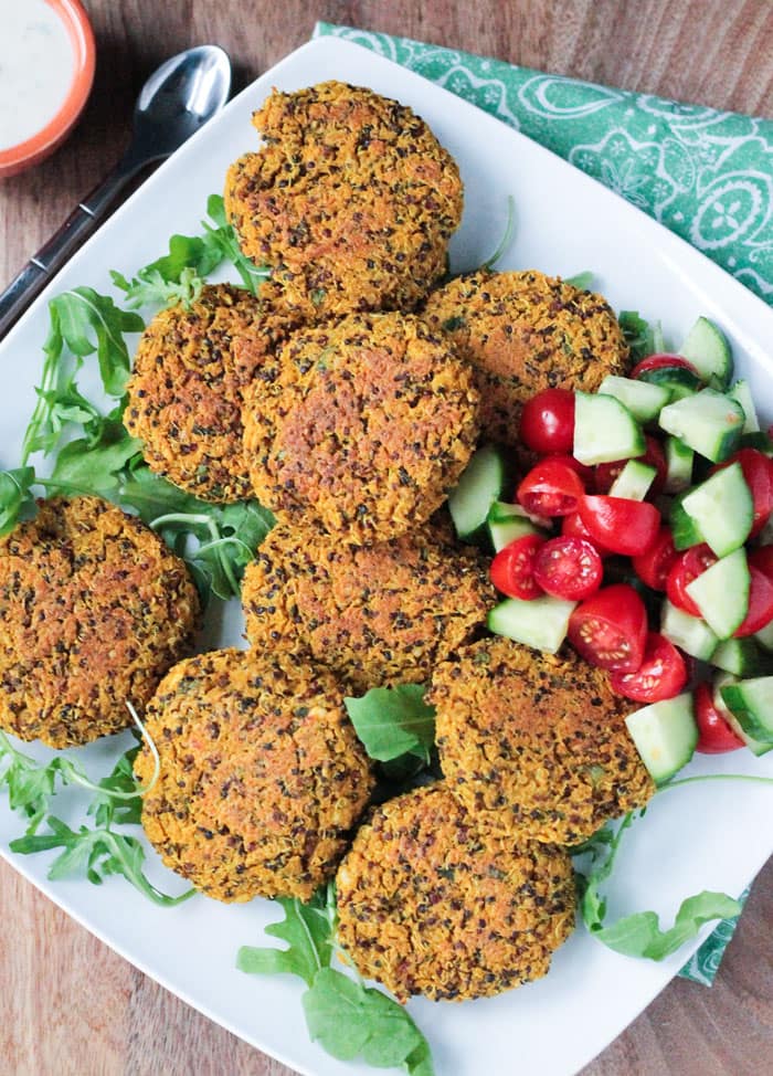 Gluten Free Red Lentil Quinoa Fritters - spiced with turmeric, cumin and a touch of cinnamon, these crispy vegan fritters are oil free and baked, not fried. An easy, high protein, meatless dinner that will delight your senses. Plate of Baked Red Lentil Quinoa Fritters over arugula and next to a cucumber tomato salad.