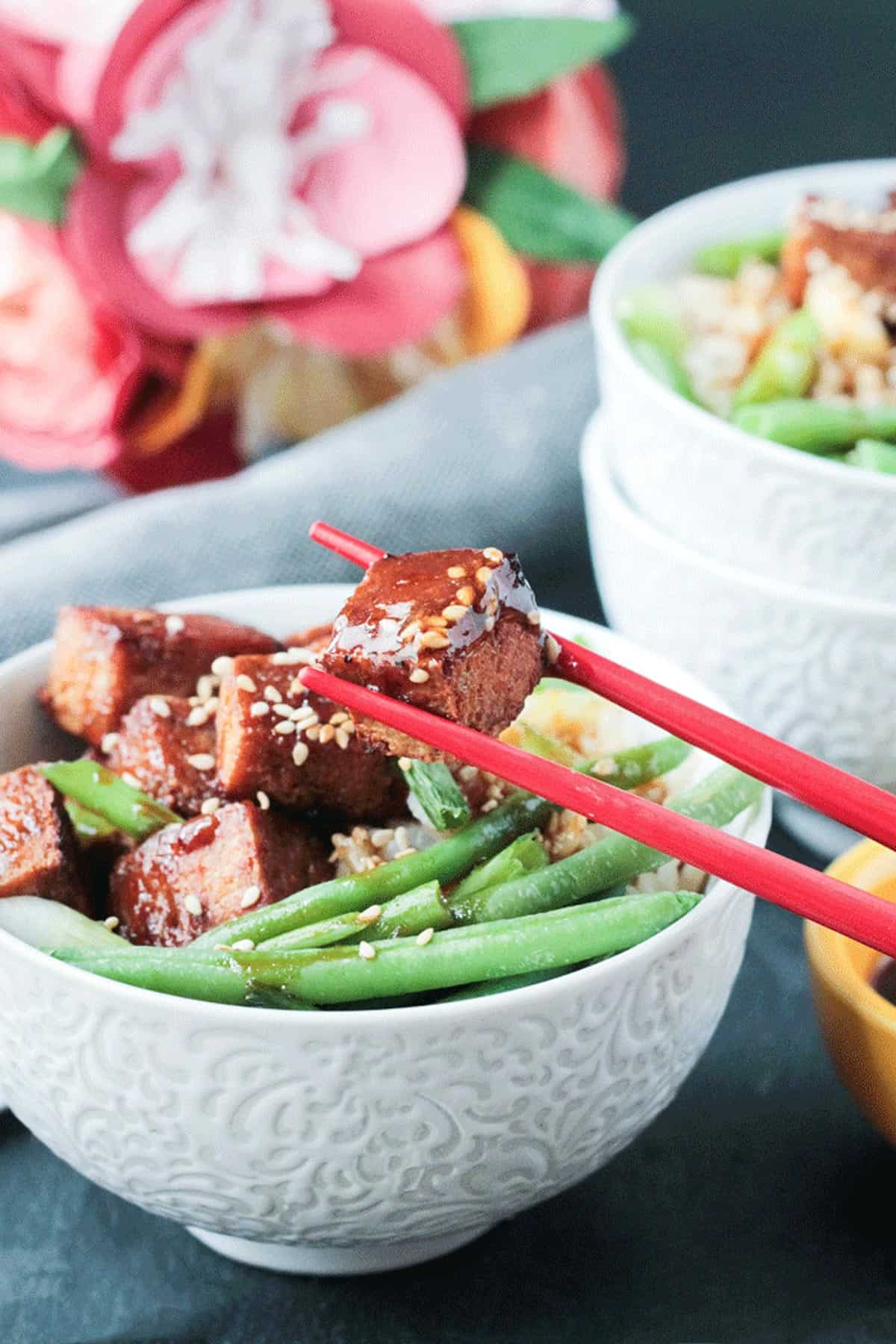 One piece of tofu being lifted out of a bowl with red chopsticks.