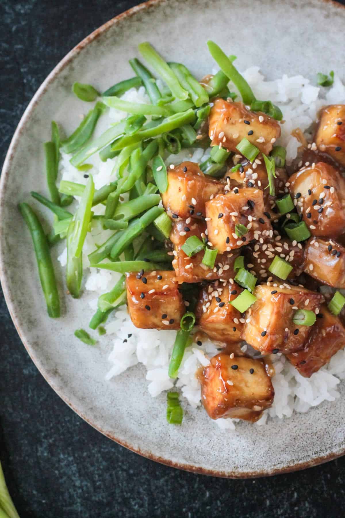 Bite size chunks of baked sesame ginger tofu over rice with green beans and garnished with scallions and sesame seeds.