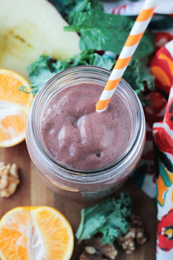Overhead view of a Superfood Smoothie in a glass jar with an orange and white striped straw. Fresh halved oranges and apples, kale leaves, and raw walnuts scattered around the jar.