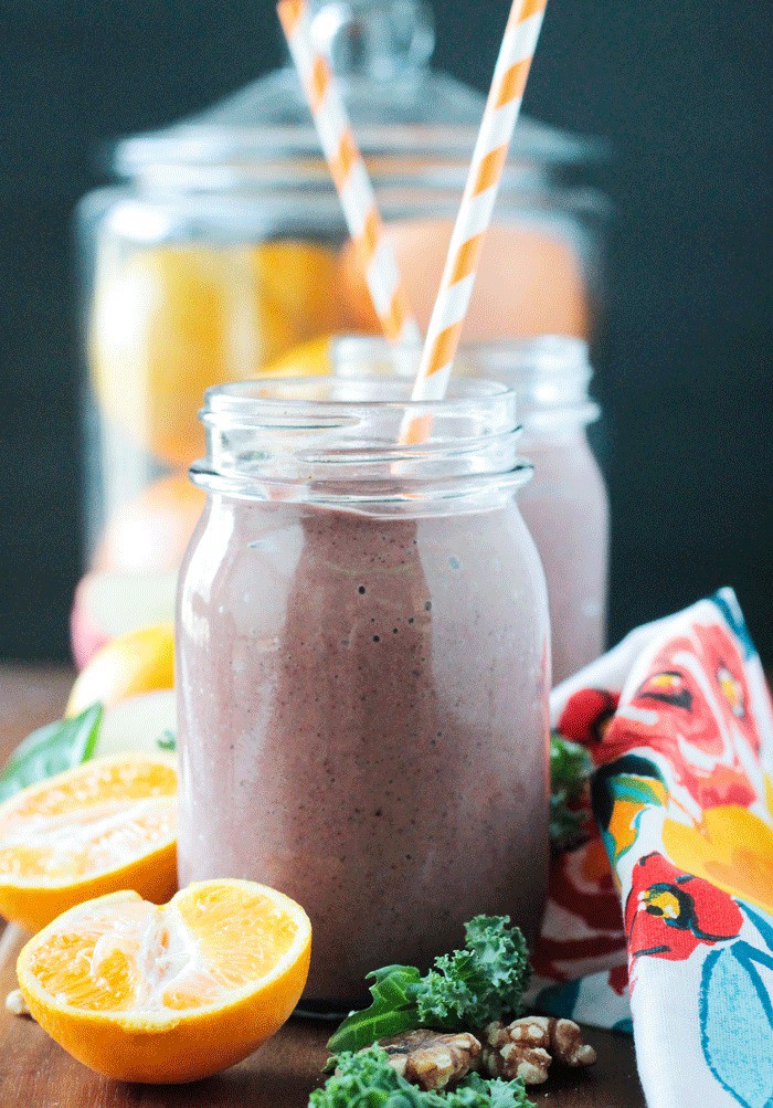 Two purple smoothies in glasses - one behind the other. Both with orange and white striped straws. Oranges halves next to the glasses. A large glass container of oranges behind both smoothies.