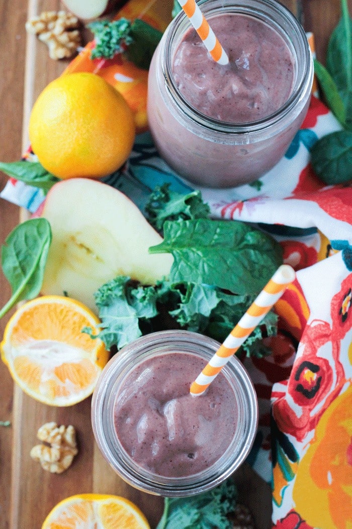 Two purple superfood smoothies in glasses with fresh fruit next to them on the table.