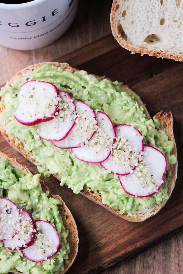 Piece of toast on a wooden serving board with smashed avocado, sliced radishes lined up across the avocado, and hemp seeds sprinkled on top of the radishes. 