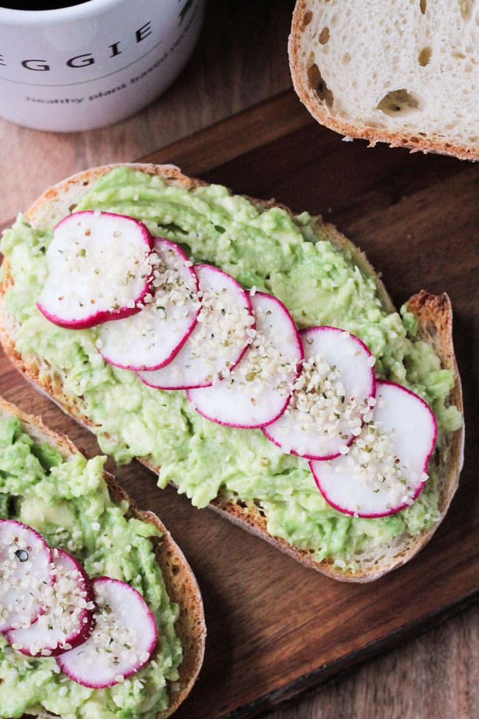 Piece of toast on a wooden serving board with smashed avocado, sliced radishes lined up across the avocado, and hemp seeds sprinkled on top of the radishes.