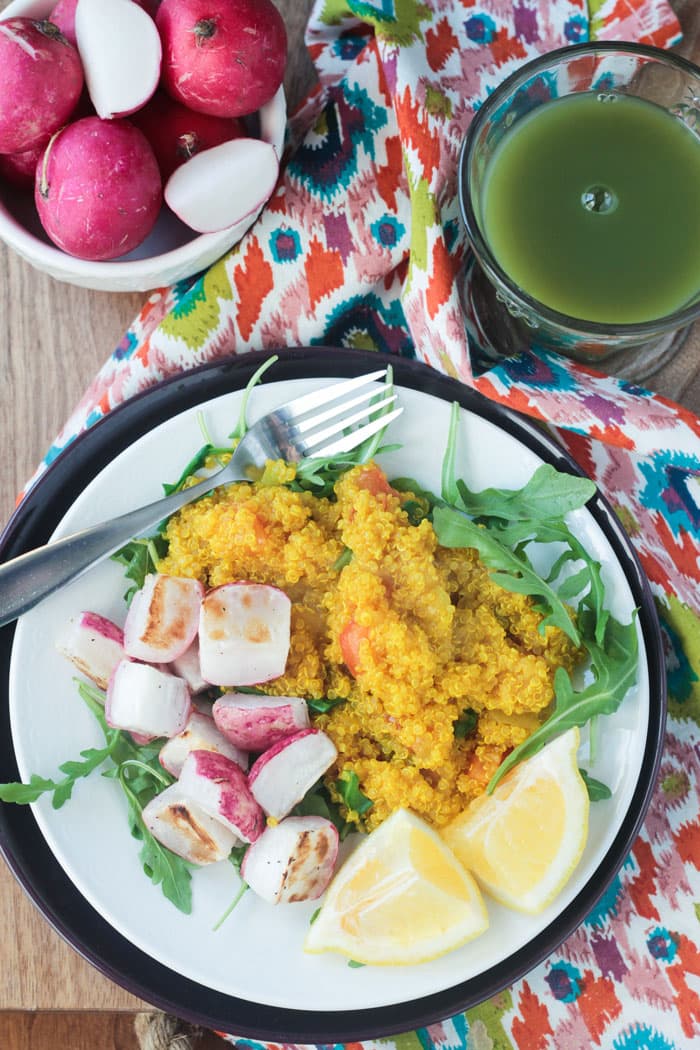 Breakfast plate of turmeric quinoa, chopped roasted radishes, arugula, and two lemon wedges. Bowl of raw radishes behind next to a glass of green juice.