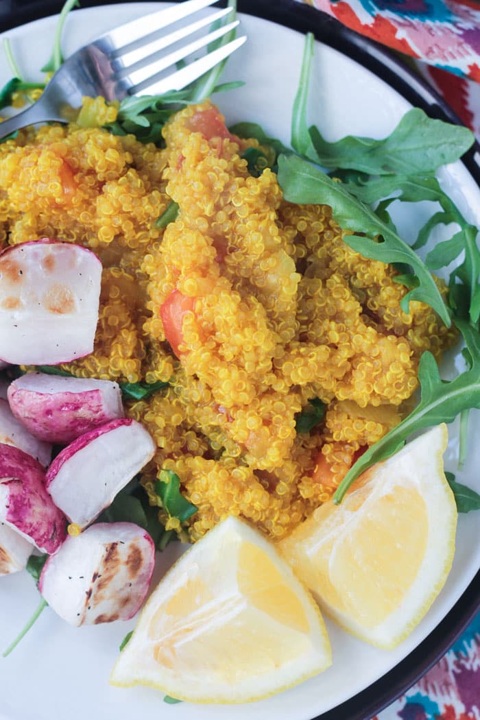 Close up overhead view of yellow hued quinoa with diced tomatoes on a plate over arugula, next to roasted radishes and lemon wedges. 