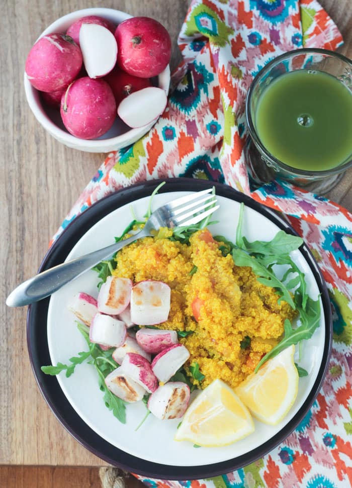 Overhead view of a plate of quinoa, radishes, arugula, and lemons in front of a bowl of radishes and glass of green juice. Brightly colored dish towel under the plate.