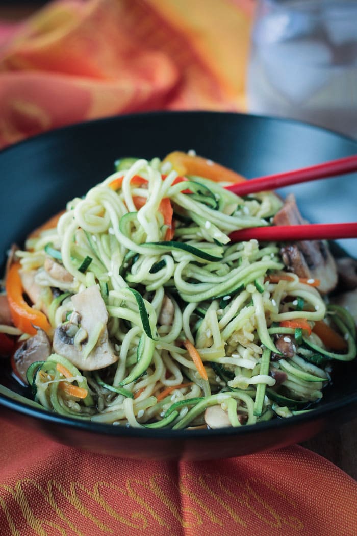 Two red chopsticks in a bowl of stir fry zucchini noodles with mushrooms and peppers.