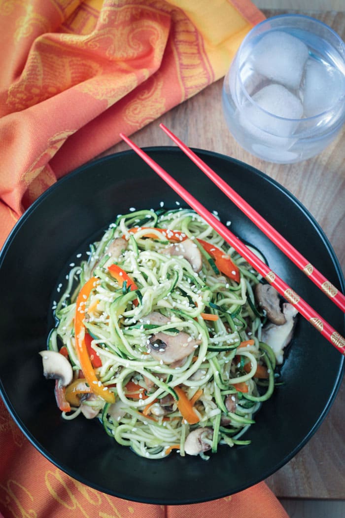 Stir Fry Zucchini Noodles in a black bowl. Two red chopsticks rest across the side of the bowl. Orange dish cloth next to the bowl and a glass of water.