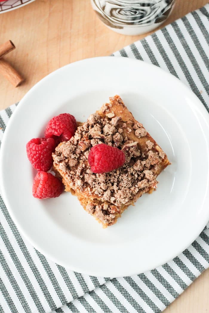 One piece of vegan cinnamon coffee cake, on a white plate, topped with one fresh raspberry. Three raspberries on the plate next to the cake.