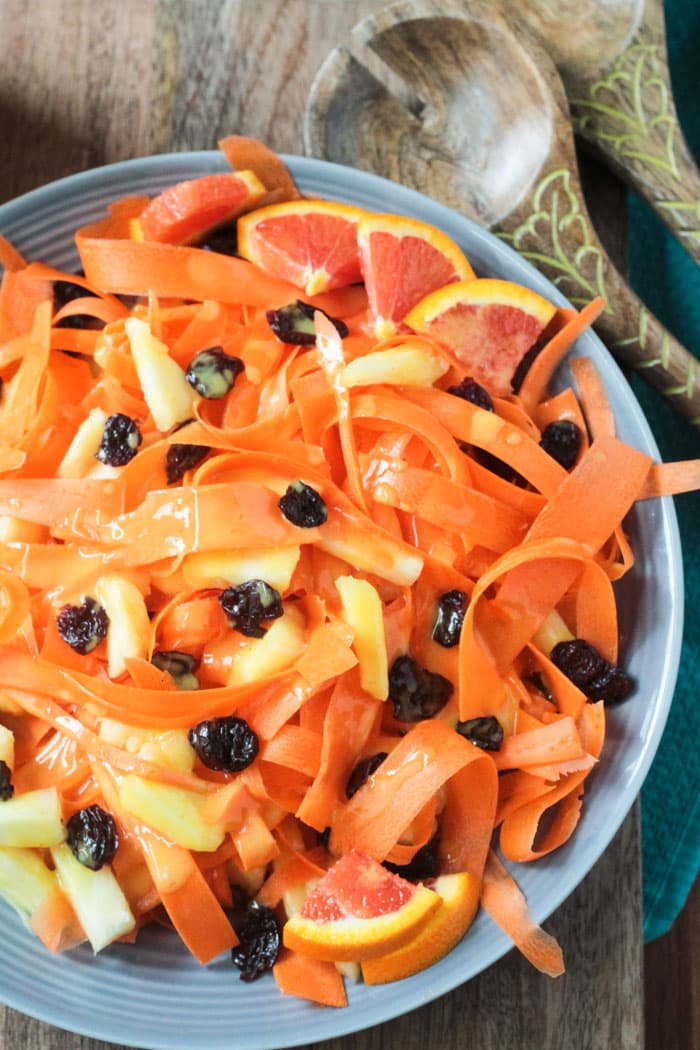 Carrot Pineapple Salad on a gray plate. Orange wedges on the side. Wooden salad serving spoons next to the plate.