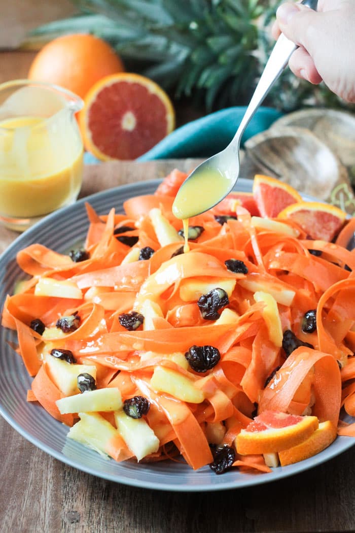 Spoon drizzling orange dressing onto a salad made of carrot ribbons, pineapple, and dried cherries.