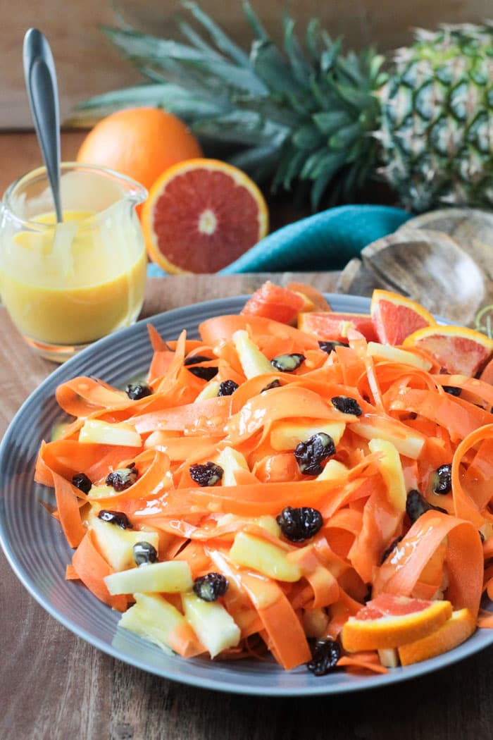 Salad of carrot ribbons, pineapple chunks, and dried cherries on a gray plate. Small jar of orange dressing with a spoon in the back ground next to fresh oranges and a whole pineapple.