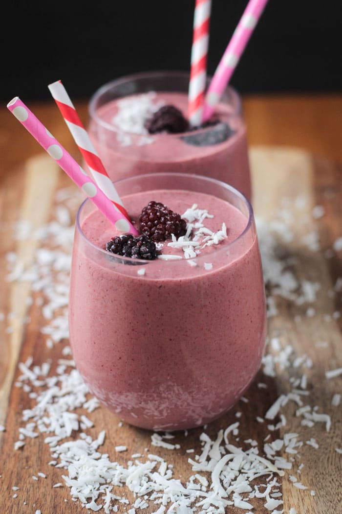 Two glasses of Coconut Blackberry Smoothie on a wooden board. Both glasses contain two straws, 2 fresh blackberries, and shredded coconut. More shredded coconut sprinkled on the board around the glasses.