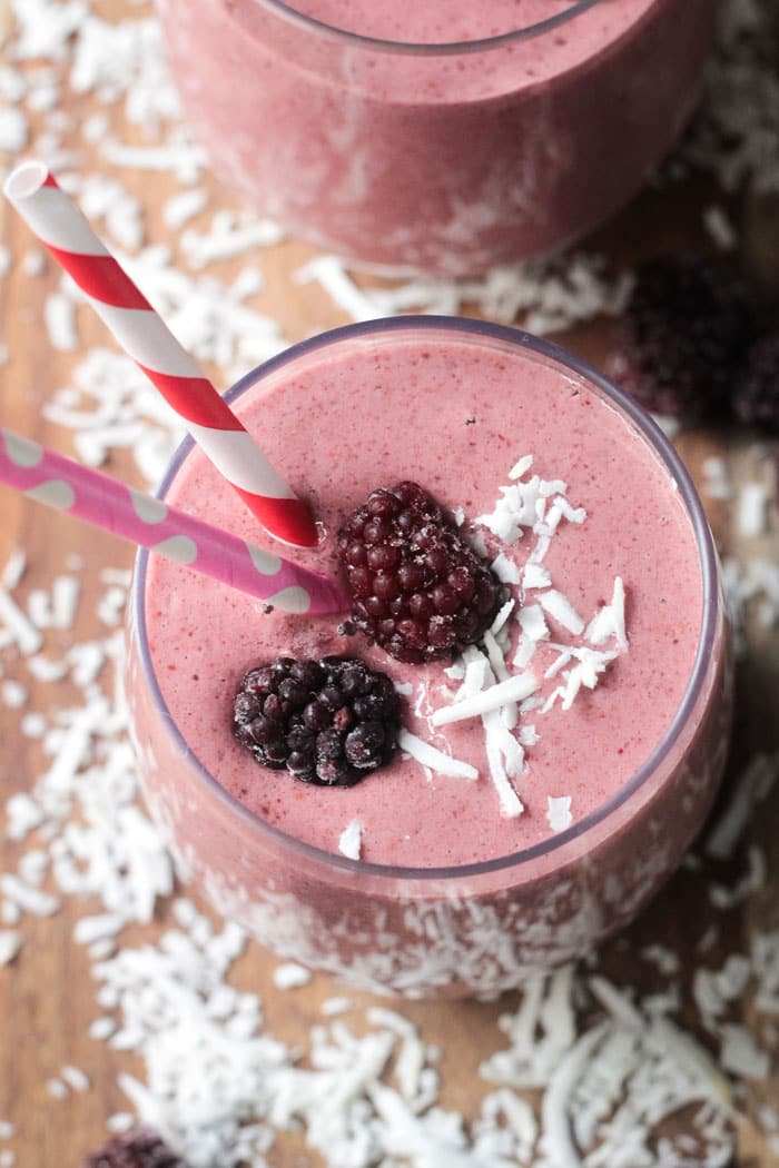 Coconut Blackberry Smoothie w/ 2 fresh blackberries and shredded coconut on top. One red and white striped straw and one pink and white polka dot straw in the glass. Shredded coconut surround the glass on the table.