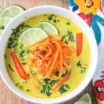 Bowl of Coconut Curry Soup with golden yellow broth, bell red peppers, spiralized sweet potato noodles, and garnished with lots of chopped cilantro and two lime wedges.