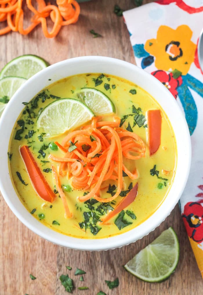 Bowl of Coconut Curry Soup with golden yellow broth, bell red peppers, spiralized sweet potato noodles, and garnished with lots of chopped cilantro and two lime wedges.