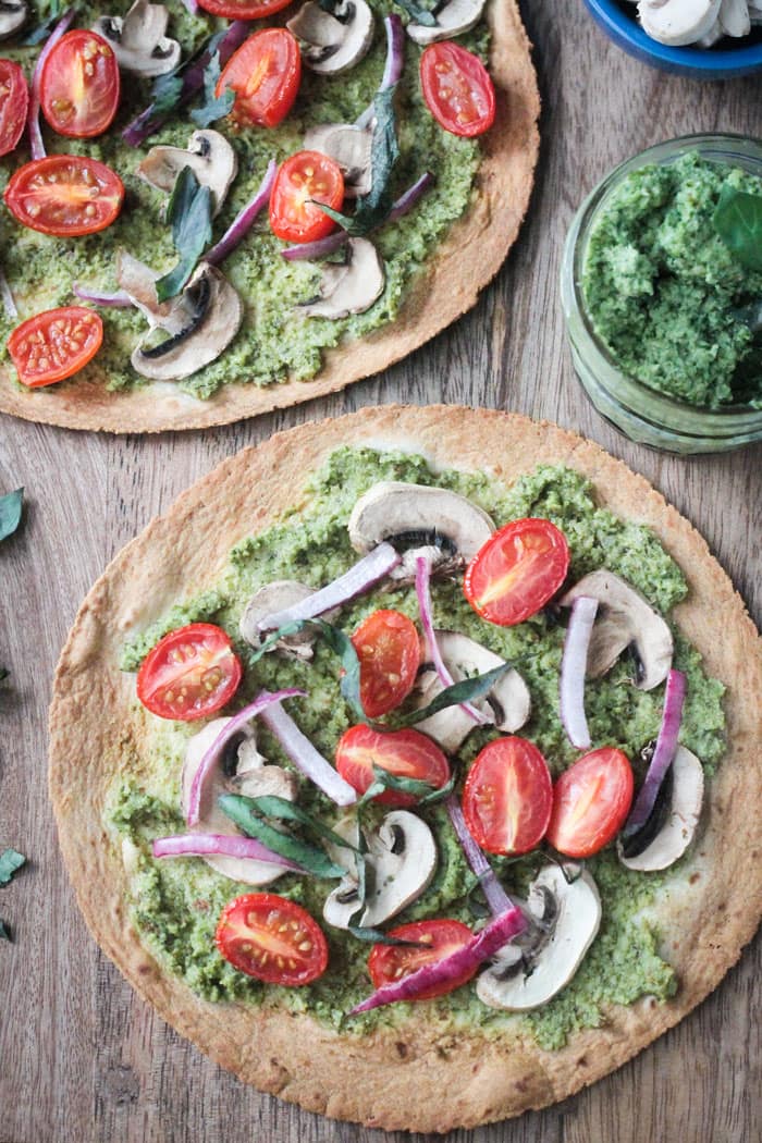 Two tortilla pizzas with pesto, cherry tomatoes, mushrooms, and red onion next to a small glass jar of pesto.