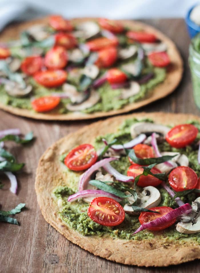 Two Vegan Tortilla Pizzas, on a wooden cutting board, topped with tomatoes, onion, mushrooms, and pesto.