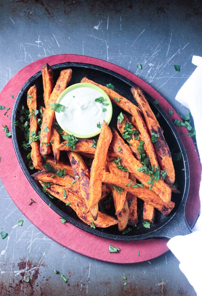 Black skillet of sweet potato fries sprinkled with parsley and a small dip bowl of white creamy sauce.