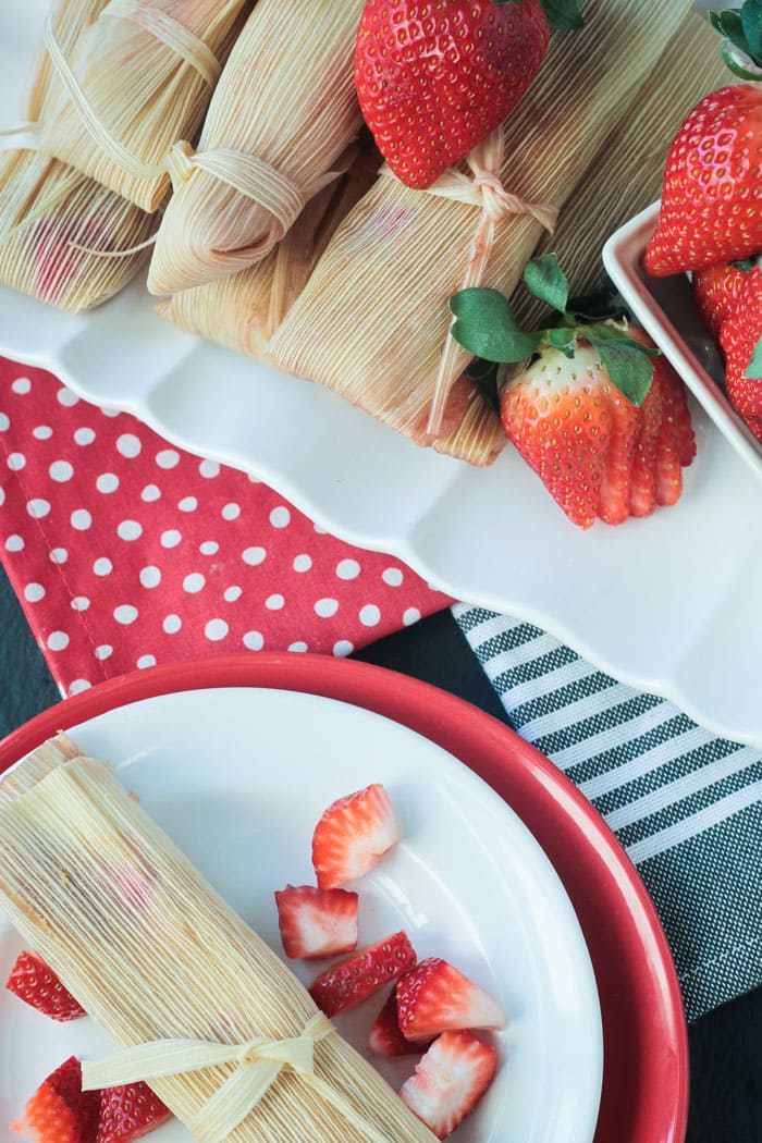 One Vegan Strawberry Dessert Tamale on a white plate with fresh cut strawberries. Plate of more tamales in the background.