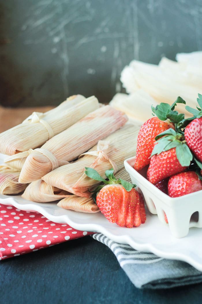 A plate of vegan tamales next to a white berry basket of fresh strawberries.