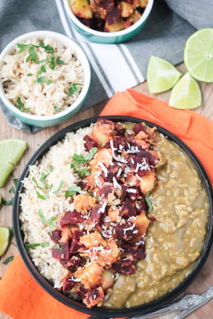 Indian dinner of rice, beets and potatoes, and lentils lined up in a black bowl. Small bowl of rice next to lime wedges in the background.