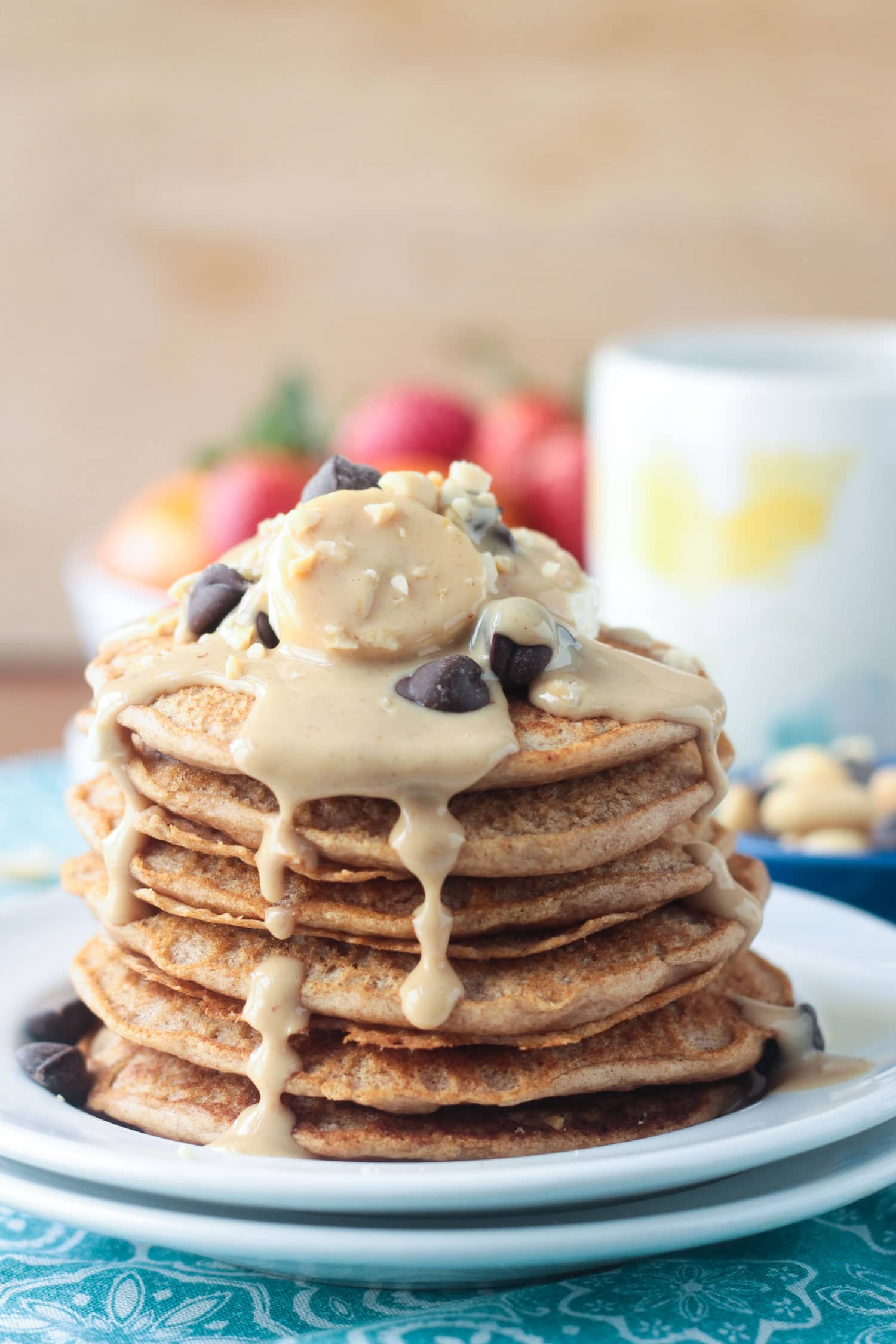 Stack of peanut butter pancakes drizzled in peanut butter syrup and topped with fresh banana slices and chocolate chips. A bowl of fresh oranges and berries and a coffee mug in the background.