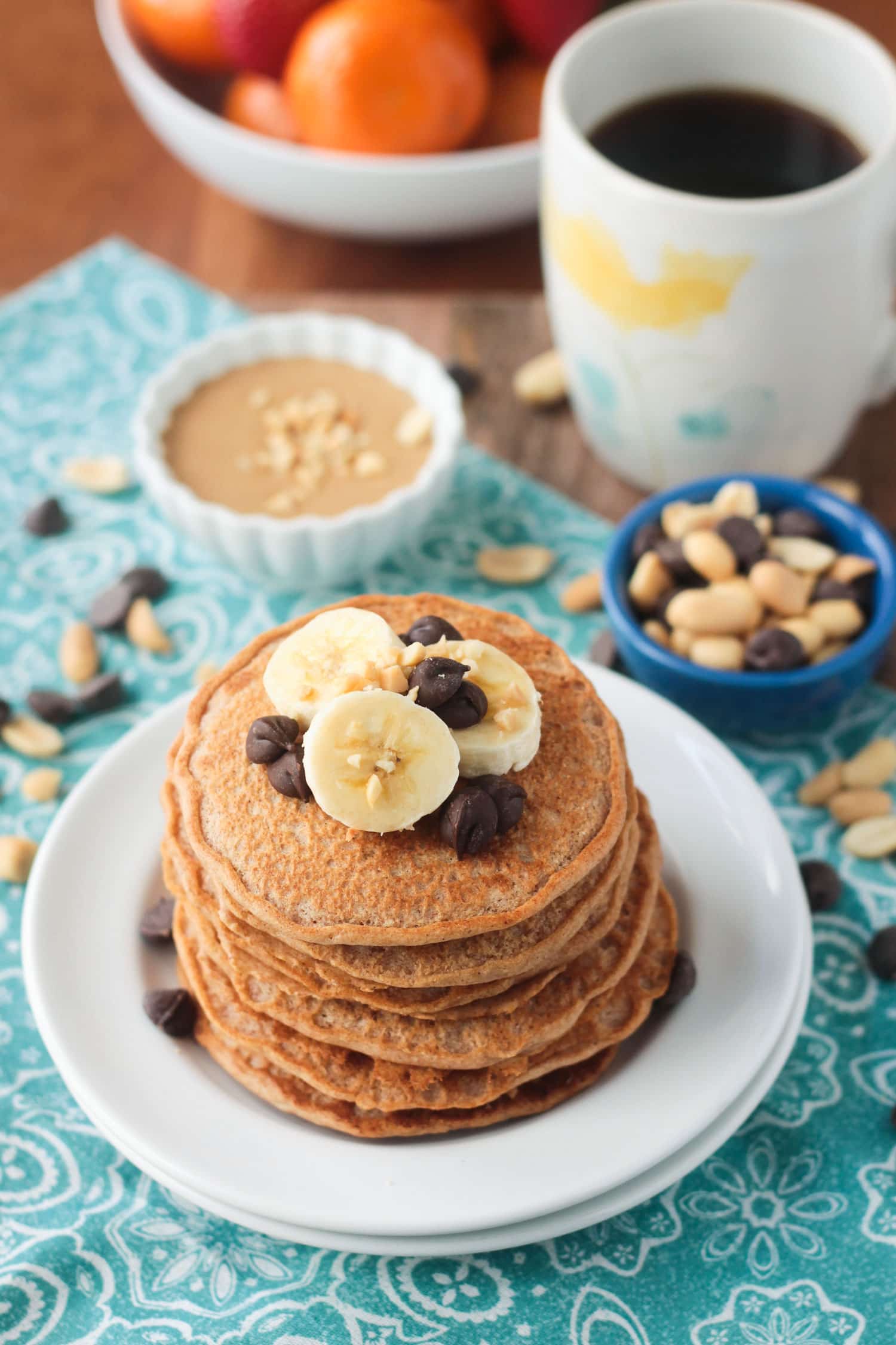 Stack of pancakes topped with banana slices and chocolate chips. Cup of coffee and a bowl of oranges in the background.