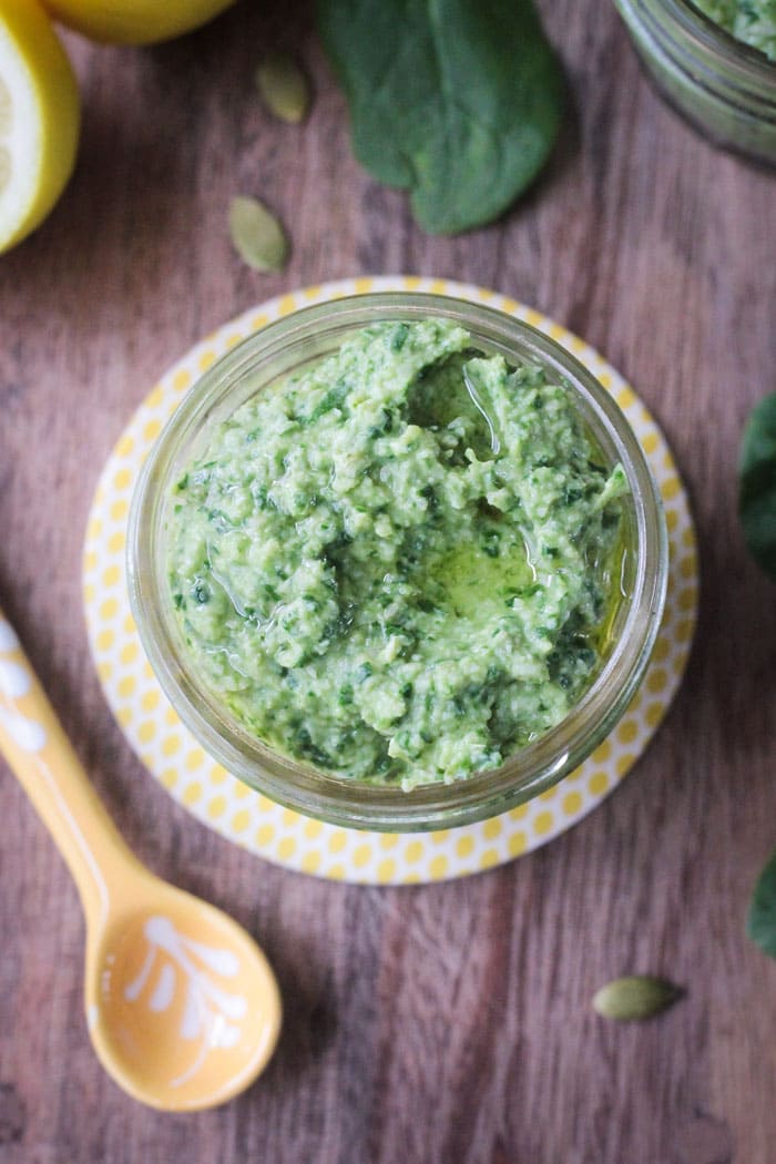 Glass jar of Spinach Artichoke Pesto topped with olive oil. Small yellow measuring spoon nearby.