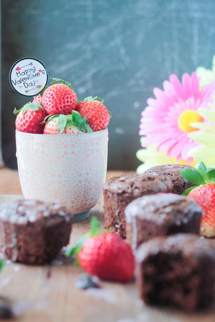 A small pink cup of fresh strawberries, with a Happy Valentine's Day cupcake topper sticking out of the top, surrounded by tahini chocolate chip cupcakes. Pink flowers in the background.