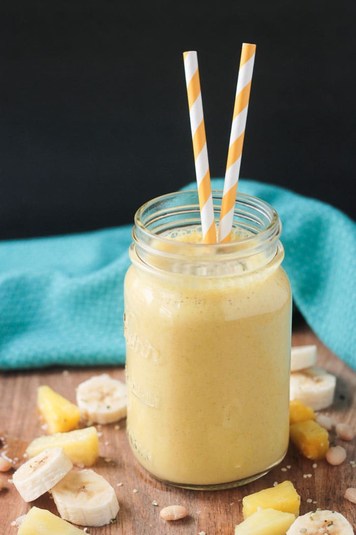 Pineapple smoothie in a mason jar with two orange/white striped straws. Banana slices and pineapple chunks around the jar. Blue dish cloth in the back.