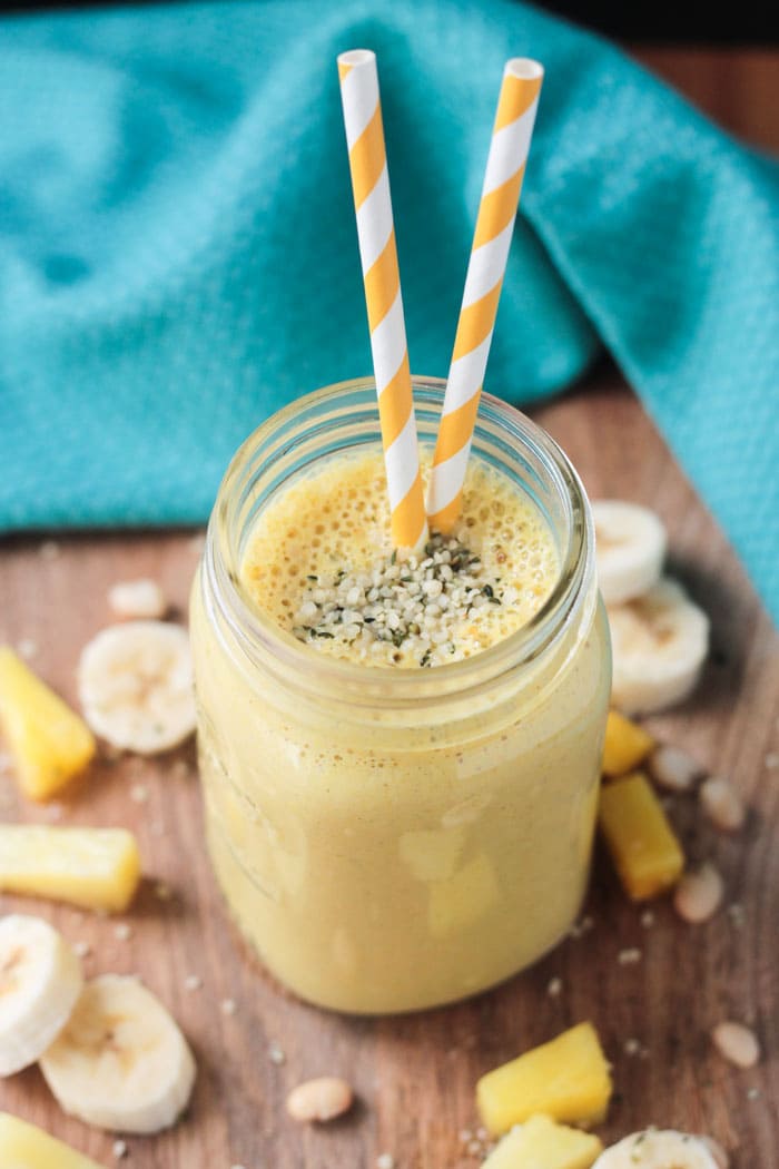 Post Workout Pineapple Smoothie topped with hemp hearts and two orange and white striped straws. A blue dish cloth in the background.