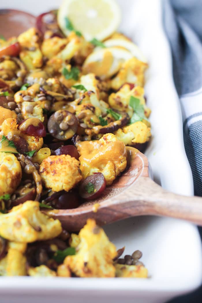 Yellow hued almond butter dressing drizzled over Roasted Cauliflower Salad. A wooden serving spoon holds a spoonful of the salad.
