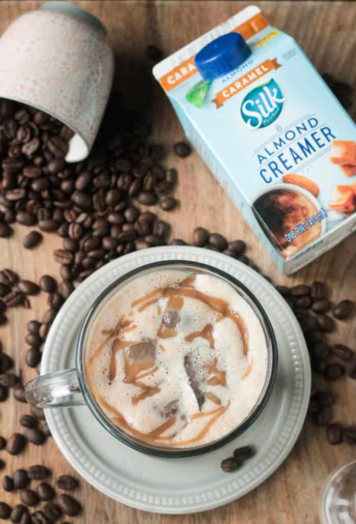 Homemade Iced Caramel Macchiato in a glass coffee mug, next to a container of Silk Almond Caramel creamer, and a cup of spilled coffee beans.