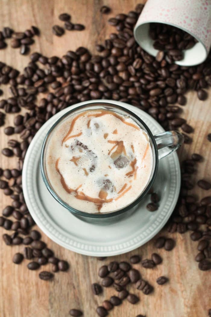 Homemade Iced Caramel Macchiato in a glass coffee mug on a small beige plate. Coffee beans strewn about around the plate.