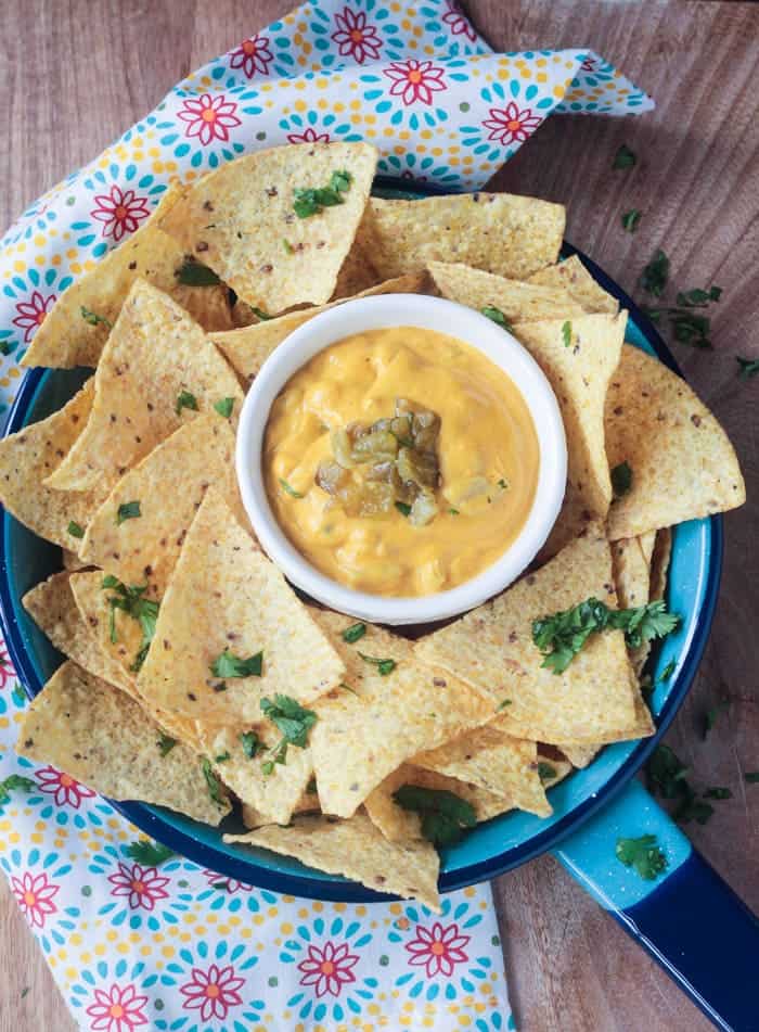 A small white bowl of Homemade Vegan Queso dip surrounded by a piles of tortilla chips sprinkled with cilantro.