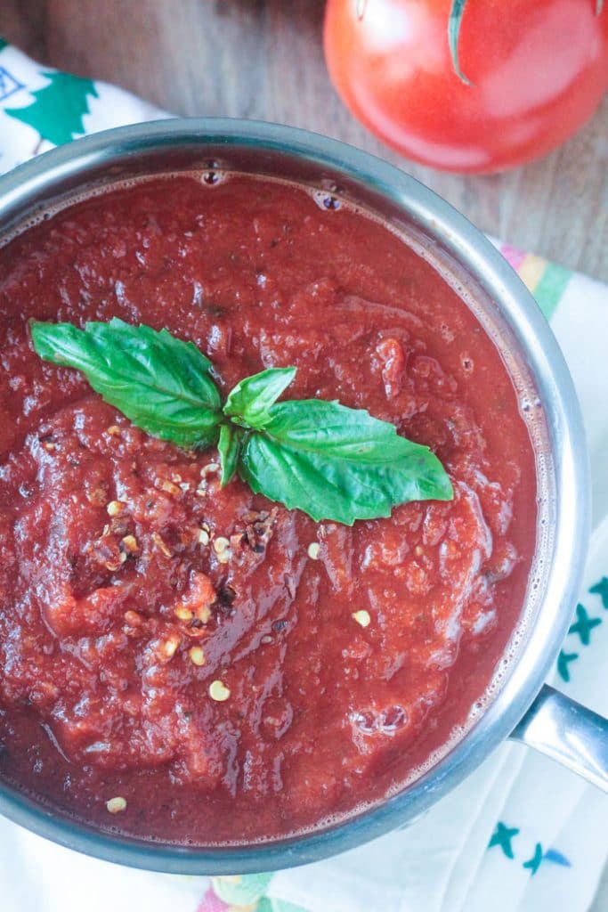 Closeup overhead image of Spicy Marinara Sauce in a pot with fresh basil leaves on top. Fresh tomatoes are sitting nearby.
