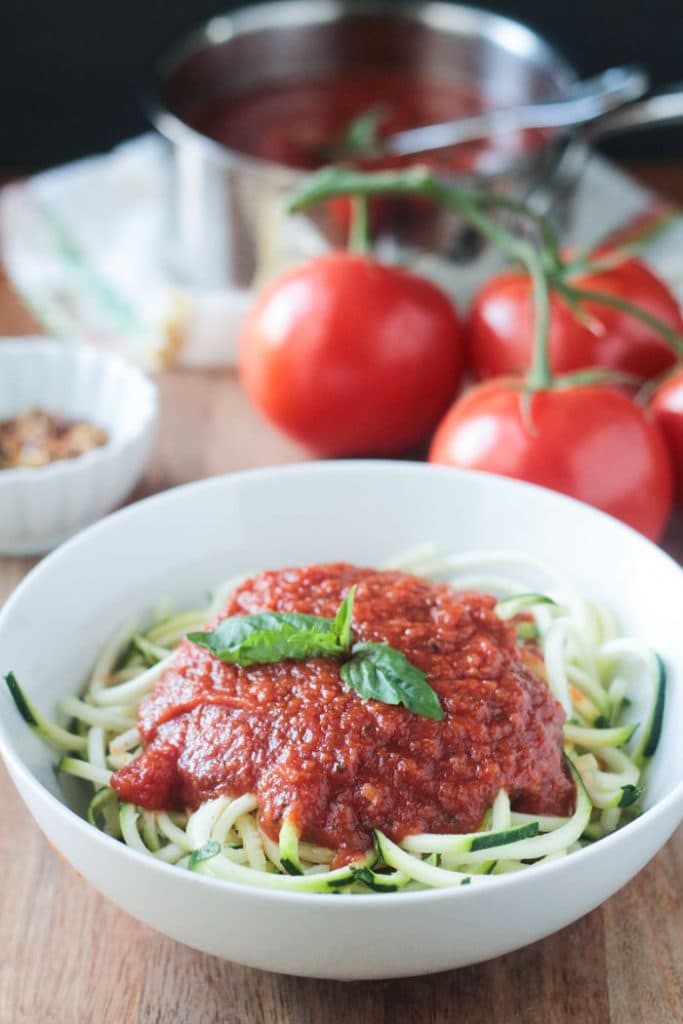 A bowl of zucchini noodles topped with Spicy Marinara Sauce and fresh basil leaves. In the background, fresh tomatoes and the of marinara sauce.