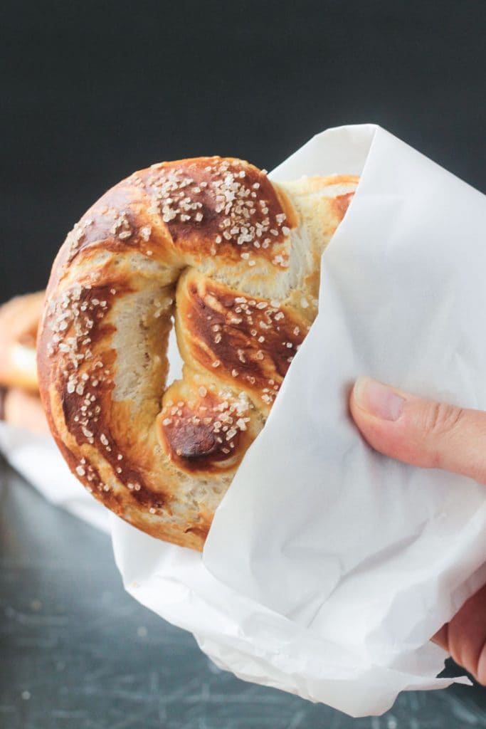 Hand holding a soft baked pretzel partially wrapped in a piece of parchment paper.