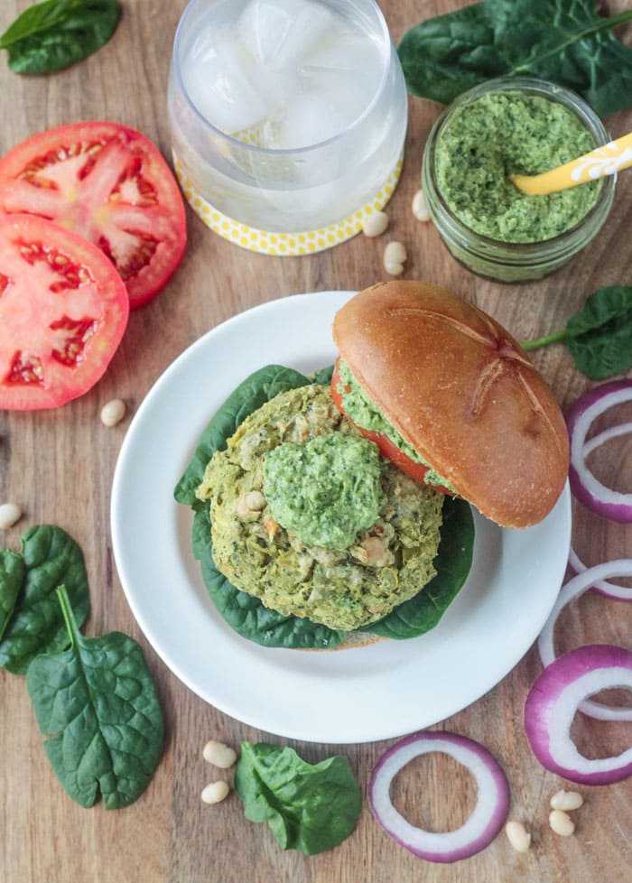 Spinach Artichoke White Bean Burger on a bottom bun with fresh spinach and topped with a dollop of pesto. The top bun is off to the side. Jar of pesto, glass of water, and two tomato slices behind the burger plate.