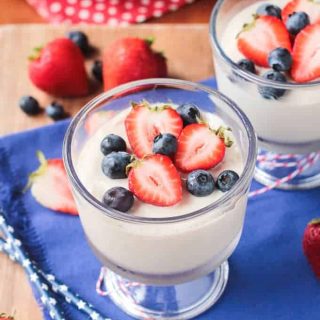 Two dessert glasses of vanilla pudding, on a blue dish towel, topped with blueberries and strawberries. More berries in the background.