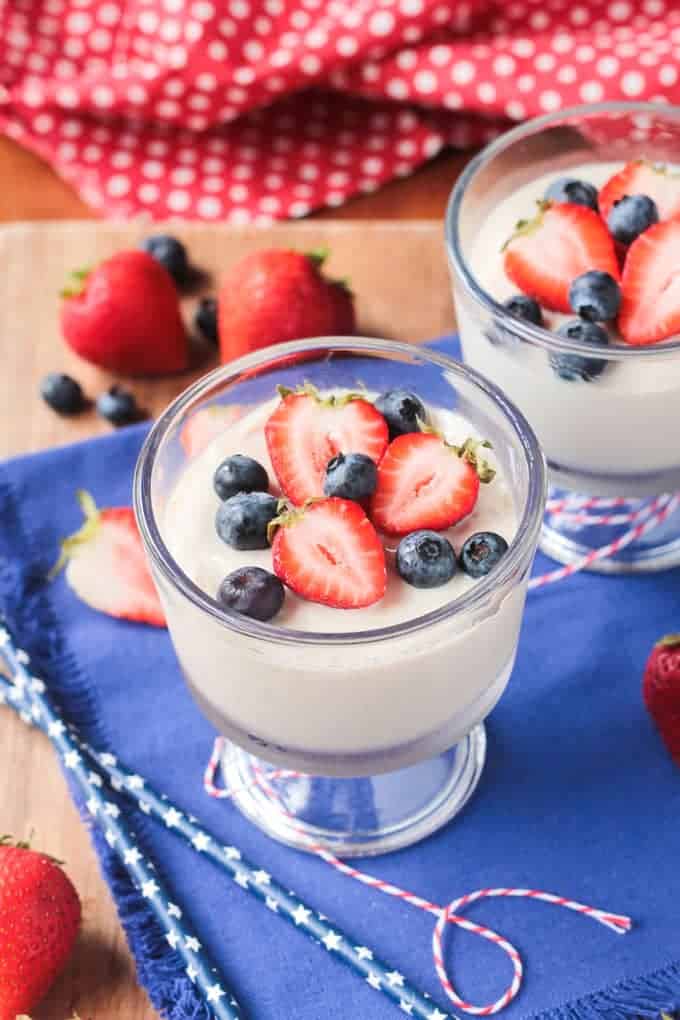 Two dessert glasses of vanilla vegan pudding, on a blue dish towel, topped with blueberries and strawberries. More berries in the background.